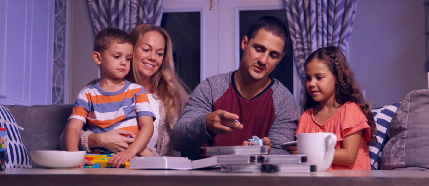 A little boys with a striped shirt sites on his mother's lap, while they watch dad assembling the Moonlite with his daughter, who has long brown hair and is in a peach color tshirt. They are all sitting on the couch