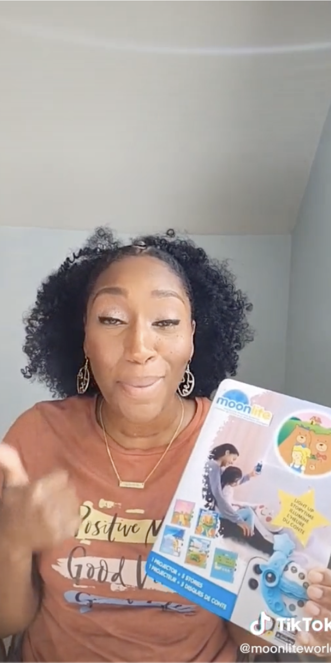 A black woman in a caramel colored tshirt holds a Moonlight box , giving a review